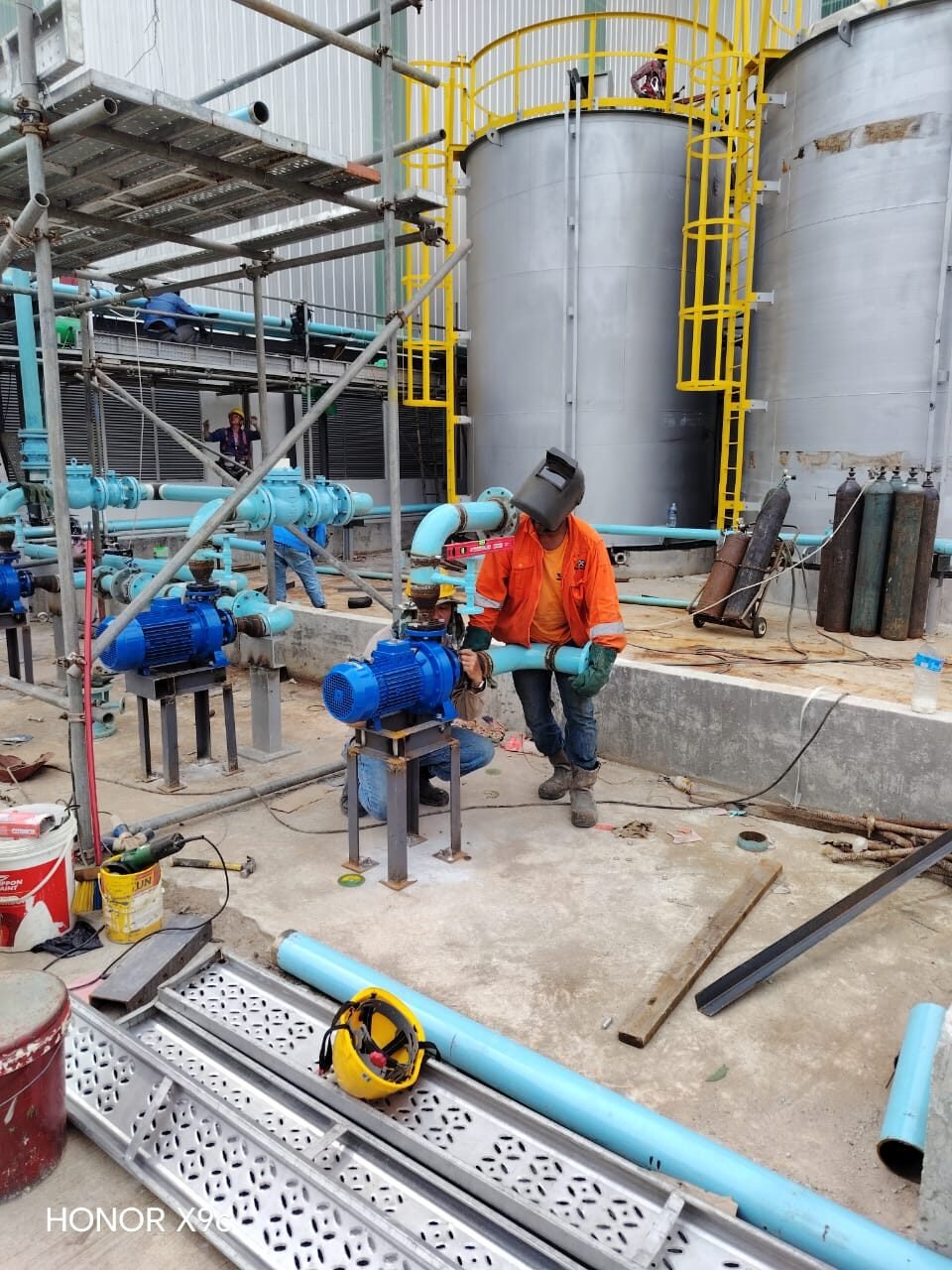 Welder connecting pipework into a pump station beside storage tanks